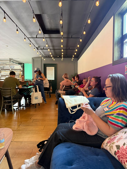A woman knitting on a blue couch during Stitch ’n Bitch, with Rachel and other makers gathered in the background at Barley Pop Brewing.