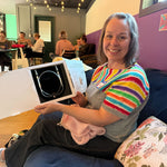 A Stitch ’n Bitch prize winner sitting on a blue couch and holding a box of circular knitting needles, with other makers gathered in the background at Barley Pop Brewing.