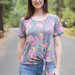 Outdoor view of the Sulky Perfect Tee sewn with a front‑tie hem in a pink and yellow floral knit, worn with light distressed jeans on a tree‑lined road