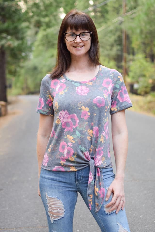 Outdoor view of the Sulky Perfect Tee sewn with a front‑tie hem in a pink and yellow floral knit, worn with light distressed jeans on a tree‑lined road