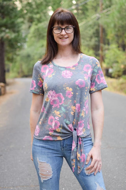 Outdoor view of the Sulky Perfect Tee sewn with a front‑tie hem in a pink and yellow floral knit, worn with light distressed jeans on a tree‑lined road