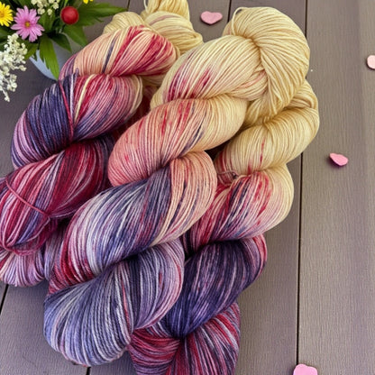 Hand‑dyed yarn in cream, pink, red, and purple tones with flowers and heart accents.