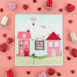 A square quilted fabric panel featuring a whimsical village scene with two pink houses, a hot air balloon, and a bird, styled on a pink background with red and silver heart-shaped confetti and spools of thread.